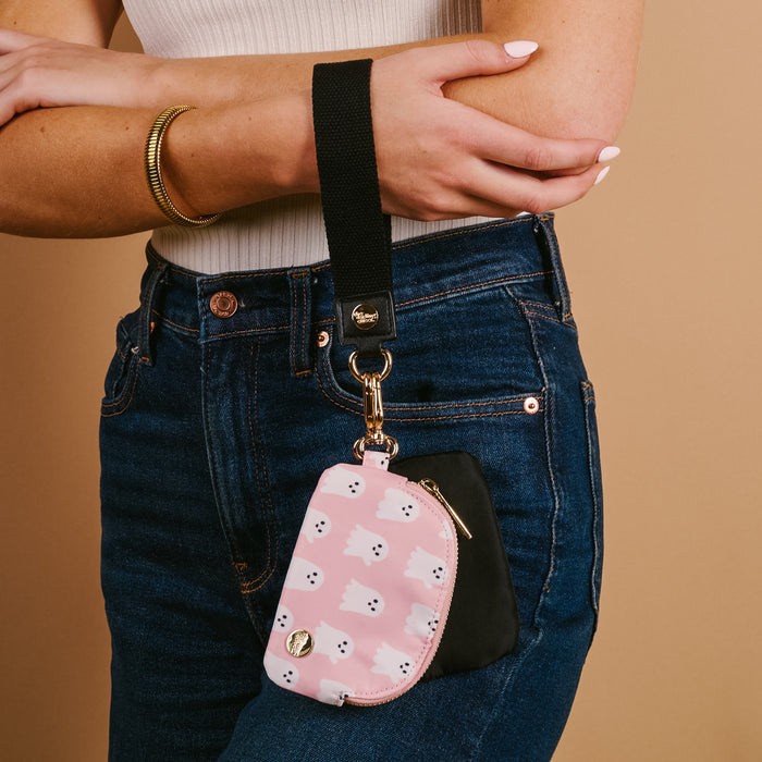 A person in blue jeans and a white top wears The Darling Effect’s Boo Crew Dynamic Duo Pouch Wristlet, a black wrist strap attached to a pink ghost-pattern pouch for carrying essentials hands-free.
