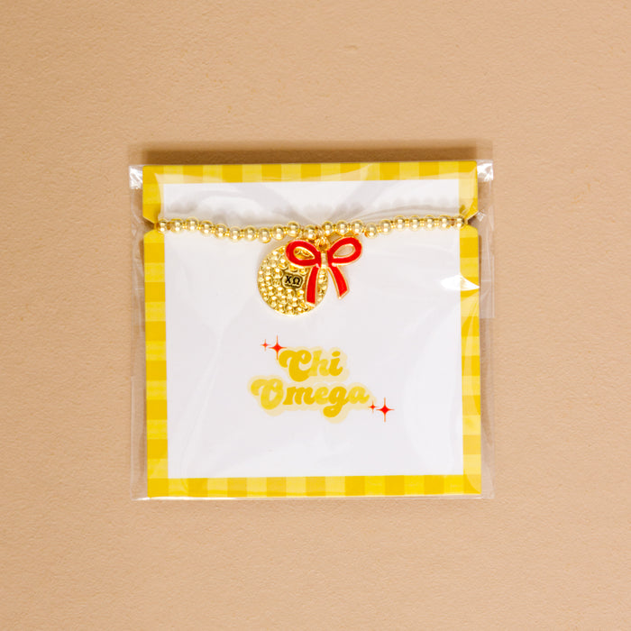The Darling Effect’s Chi Omega Bow Gold Charm Bracelet features gold beads, a round charm, and red bow on yellow gingham backing with “Chi Omega” in cursive—perfect for showing chapter pride.