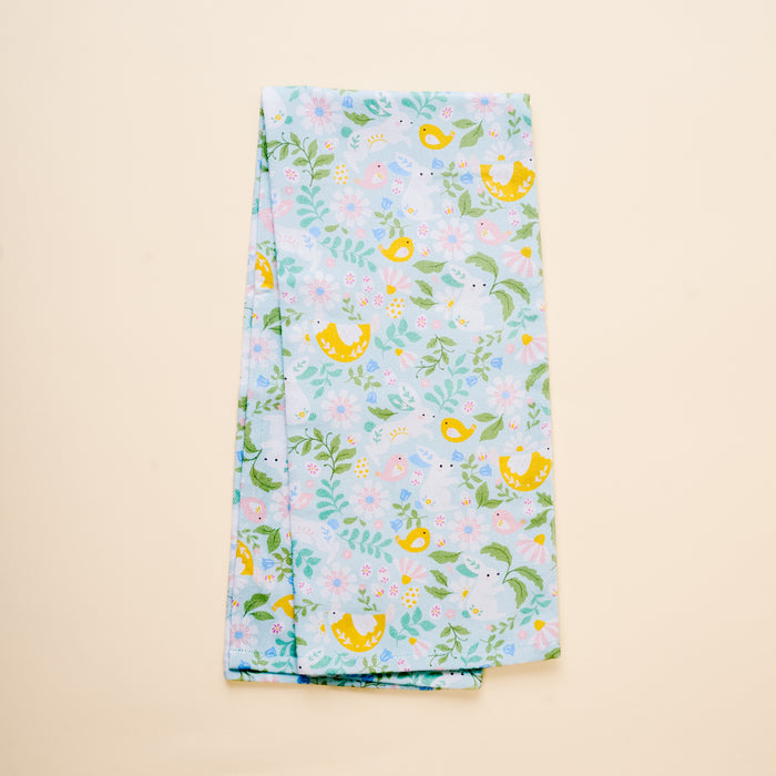 The Hoppy Days Tea Towel by the-darling-effect features yellow birds, pink and blue flowers, and green leaves on a light blue cotton background, shown folded on a plain beige surface.