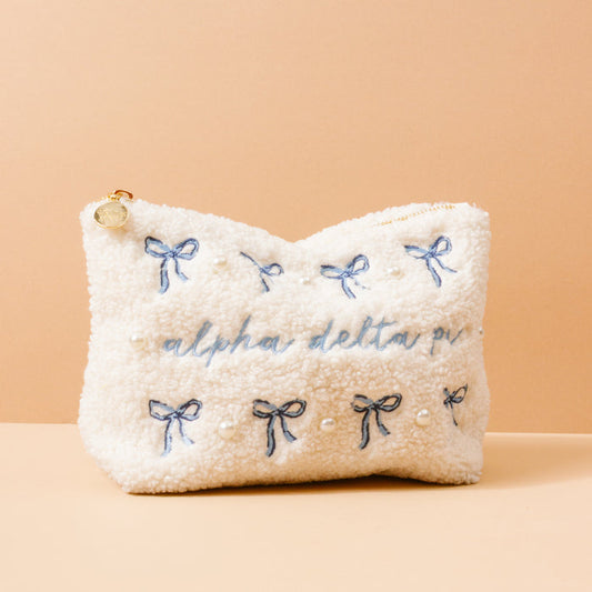 The Darling Effect Alpha Delta Pi Bows & Pearls Teddy Pouch features cream sherpa fabric, blue embroidered bows, pearl accents, and "alpha delta pi" script—making it a chic sorority gift.