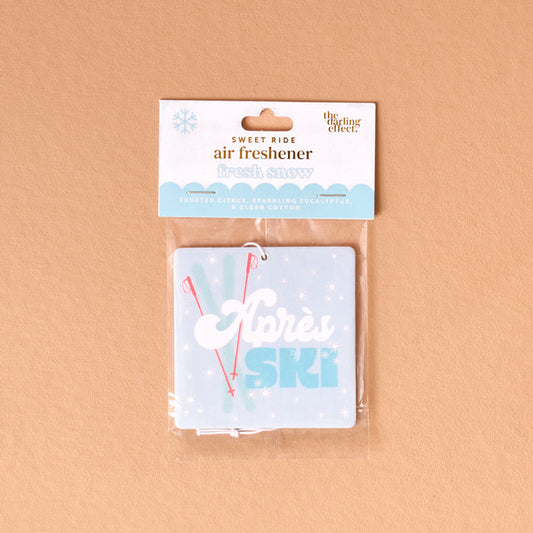 The Darling Effect’s Aprés Ski Sweet Ride Car Air Freshener features red skis and poles on a light blue background, offering a long-lasting frosted citrus, sparkling eucalyptus, and clean cotton scent for your car.