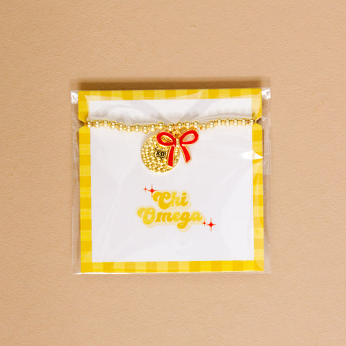 The Darling Effect’s Chi Omega Bow Gold Charm Bracelet features gold beads, a round charm, and red bow on yellow gingham backing with “Chi Omega” in cursive—perfect for showing chapter pride.