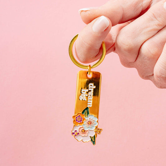 A hand holds The Darling Effect’s Dream Big Enamel Floral Keychain, featuring floral details and “dream big” engraving, against a pink background.