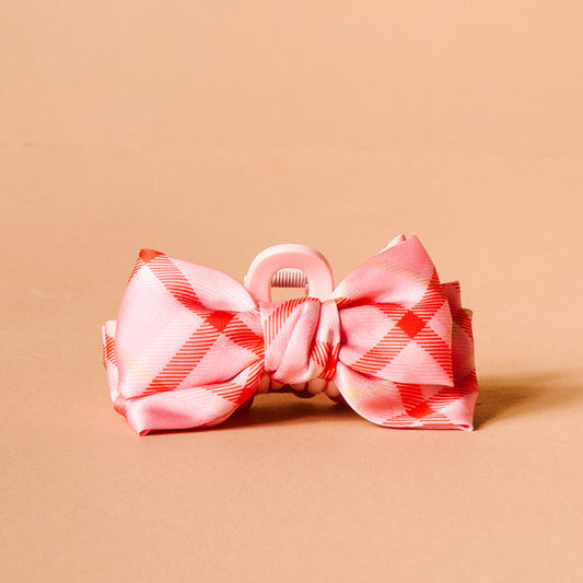 The Darling Effect's Festive Plaid Mini Satin Bow Claw Clip features a pink and red plaid satin bow on a light pink claw clip, shown on a solid beige background.