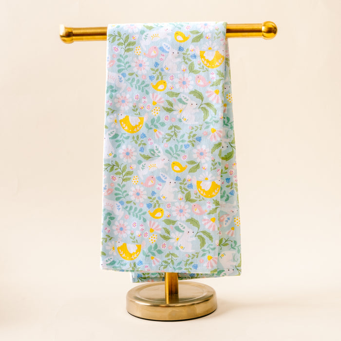 The Hoppy Days Tea Towel by the-darling-effect, featuring a vibrant pattern of yellow birds, flowers, and leaves on light blue fabric, is displayed neatly on a gold metal stand against a neutral background.