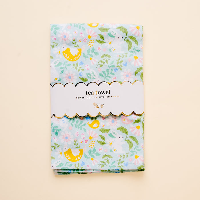 The Hoppy Days Tea Towel by the-darling-effect features a light blue floral and lemon design. It’s folded with a white scalloped label reading "tea towel" and "100% cotton kitchen towel," shown against a cream background.