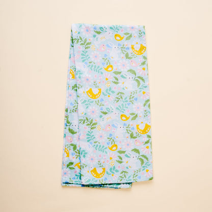 The Hoppy Days Tea Towel by the-darling-effect features yellow birds, pink and blue flowers, and green leaves on a light blue cotton background, shown folded on a plain beige surface.
