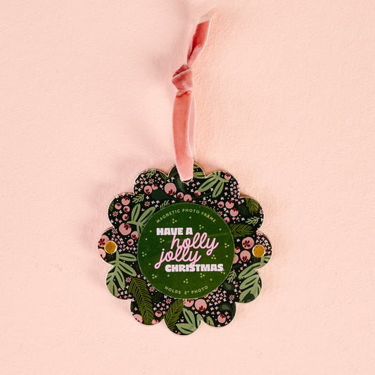 A Jolly Sprig Scalloped Round Picture Frame Ornament by The Darling Effect hangs from a pink velvet ribbon. The floral-patterned frame reads “Have a holly jolly Christmas” and holds 2 photos.